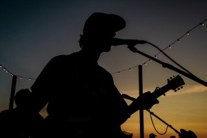 Silhouette of a musician with a guitar singing into a microphone at sunset, string lights above.