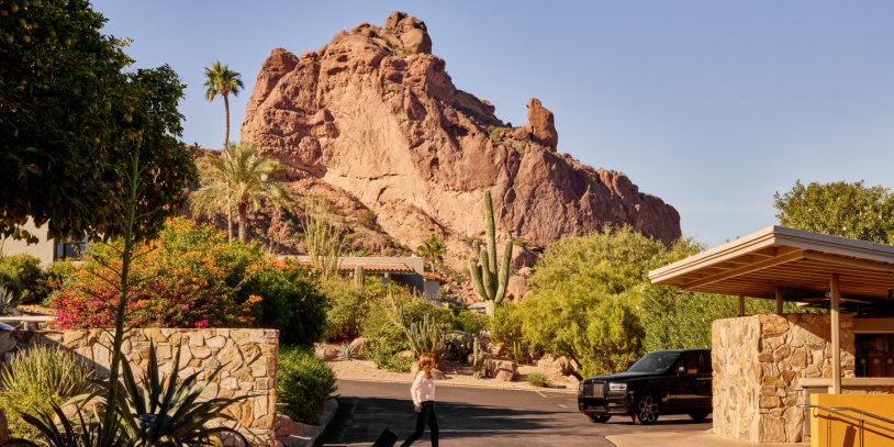 Desert scene with a rocky mountain, cactus, and greenery. A person walks a dog down a path next to a parked car under clear blue skies.