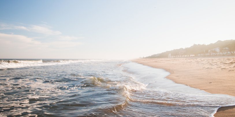Sunny beach with gentle waves, golden sand, and a clear blue sky. Coastal line fades into the distance on the right.