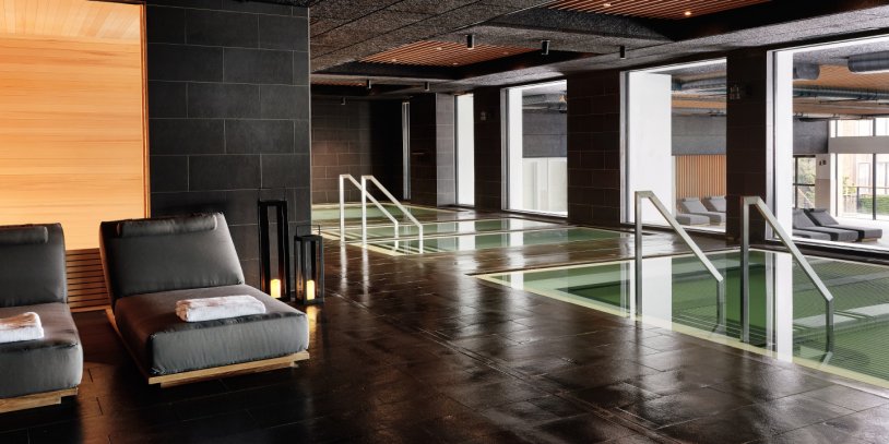 Luxurious indoor spa with two loungers, wooden accents, and a calm pool reflecting natural light from large windows.