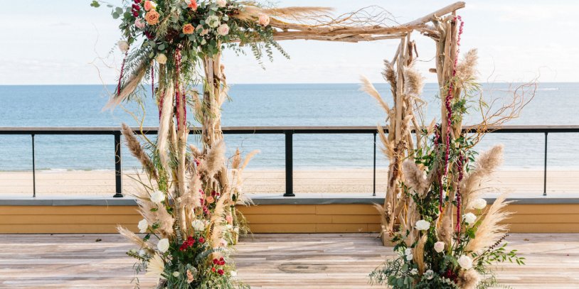Wedding arch with flowers and dried grass on a deck overlooking the beach and sea.