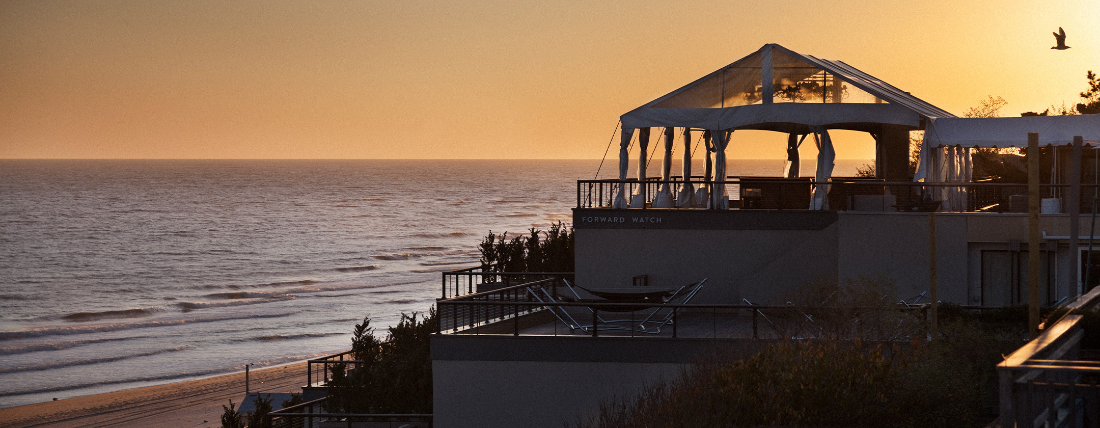 Seaside villa at sunset with a terrace and ocean view, warm orange and pink sky.