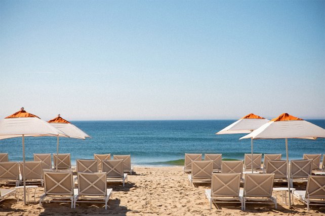 Sunlit beach with rows of white lounge chairs and umbrellas facing a calm blue sea under a clear sky.