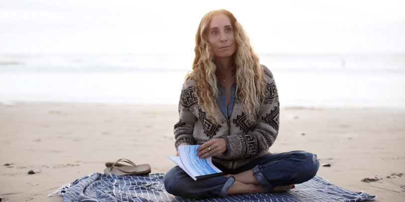 Person with long hair sitting cross-legged on a beach, holding a book, wearing a patterned sweater. Overcast sky and sea in the background.