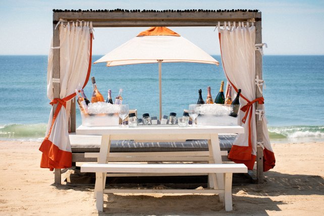 Beach cabana with a white table, drinks on ice, under a parasol, facing the ocean. Curtains tied with orange accents.