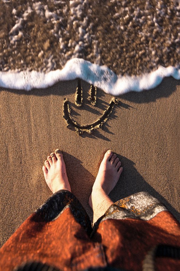 Feet on sand near a drawn smiley face, waves approaching.