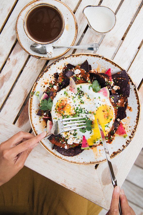 A person eats a colorful breakfast of fried eggs and vegetables at a wooden table, with a cup of coffee and cream pitcher nearby.