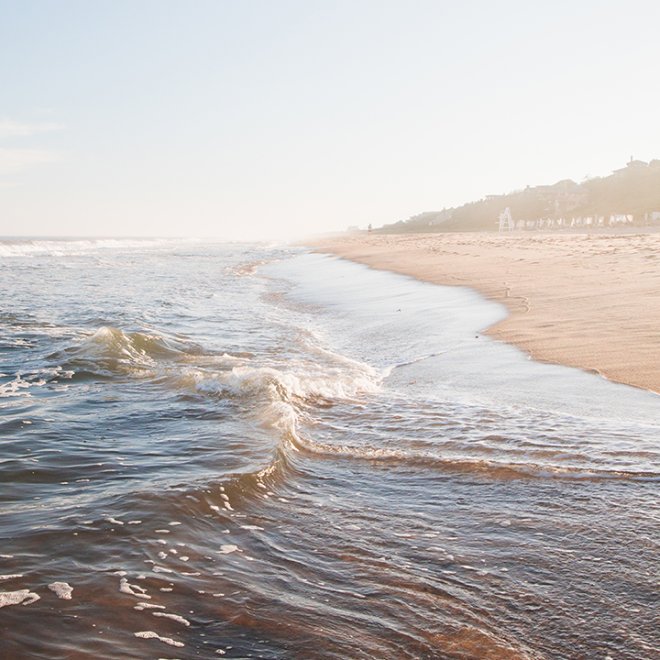 Waves gently lapping onto a sandy beach under a clear sky, with a distant view of hills and scattered buildings along the shoreline.