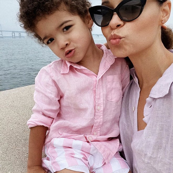 Woman and child in pink outfits pose near water, making playful kiss faces. Woman wears sunglasses. Overcast skies in the background.