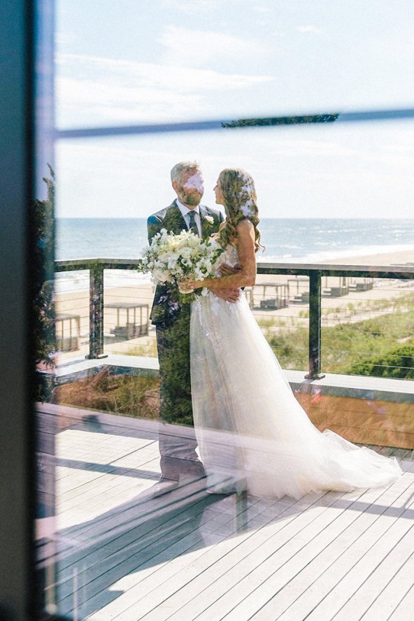 Couple in wedding attire holding flowers on a deck overlooking a beach and ocean, viewed through a window.