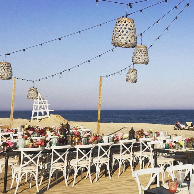 Wedding reception setup on a beach with floral arrangements on tables, white chairs, and hanging lanterns.