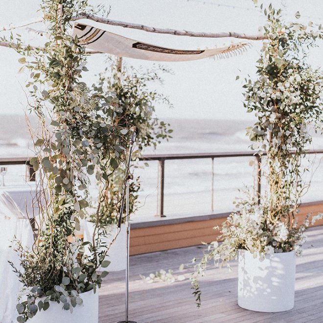 A beachside wedding altar with leafy decorations under a canopy, overlooking the ocean.