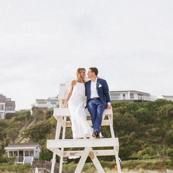 A couple in wedding attire sits on a lifeguard chair overlooking a beach, with houses and greenery in the background.