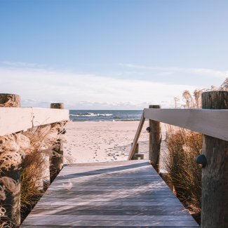 Wooden pathway leading to a sandy beach under a clear blue sky, with gentle waves visible in the background.