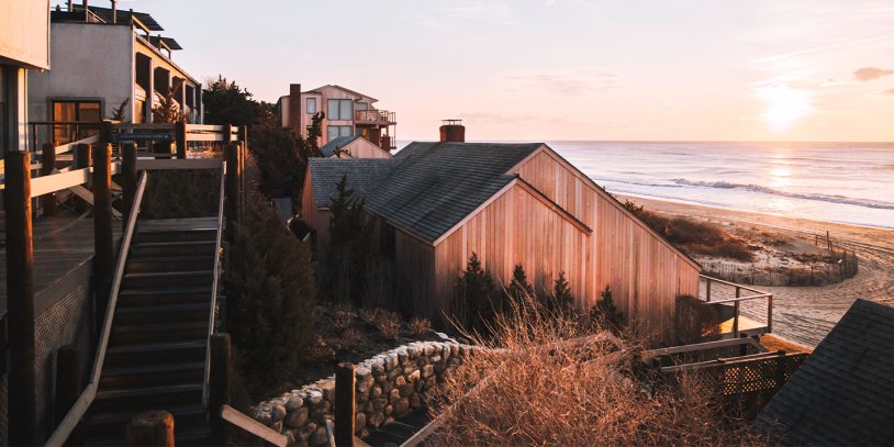 Coastal houses with wooden siding overlook the ocean at sunset, surrounded by dry grasses and a wooden staircase leading down to the beach.