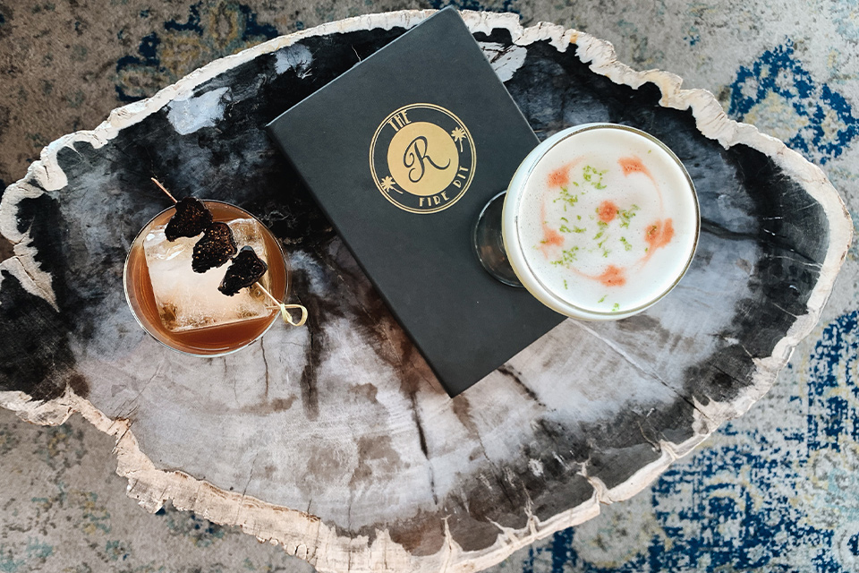 Two cocktails on a marble table with a black menu featuring a gold logo.