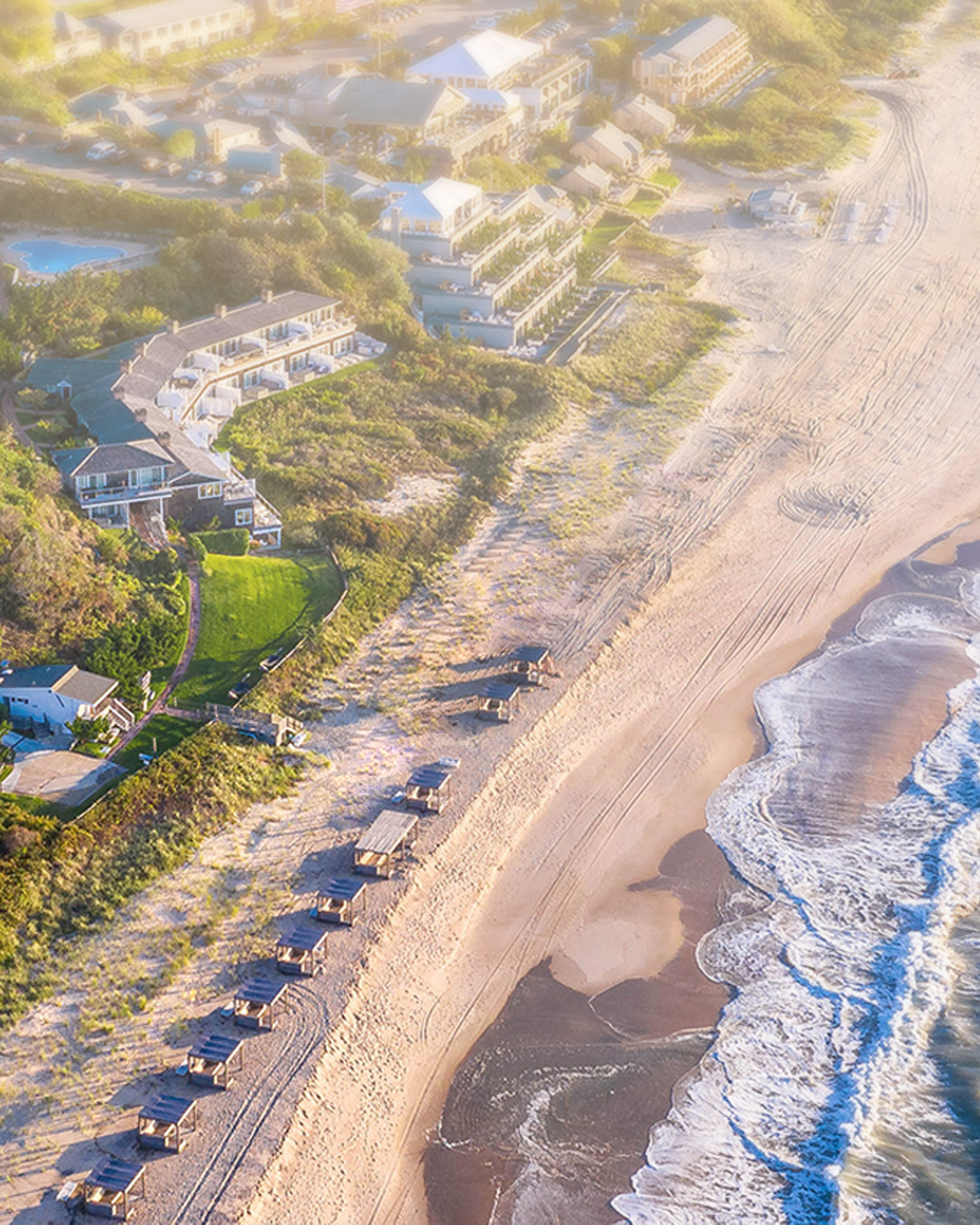 Aerial view of a coastal resort with lush greenery, buildings, and pathways alongside a sandy beach and calm ocean waves under a clear sky.