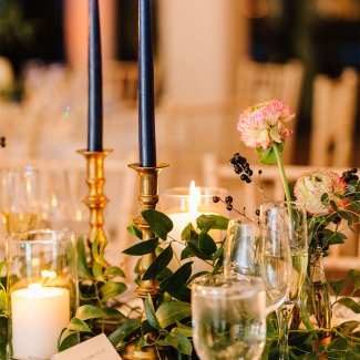 Elegant table setting with tall blue candles, pink flowers, greenery, and glassware adorned with flickering candlelight.