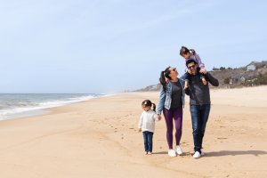 Family walking along a sandy beach, with the ocean on their left. The man carries a child on his shoulders, while another child walks beside the woman.