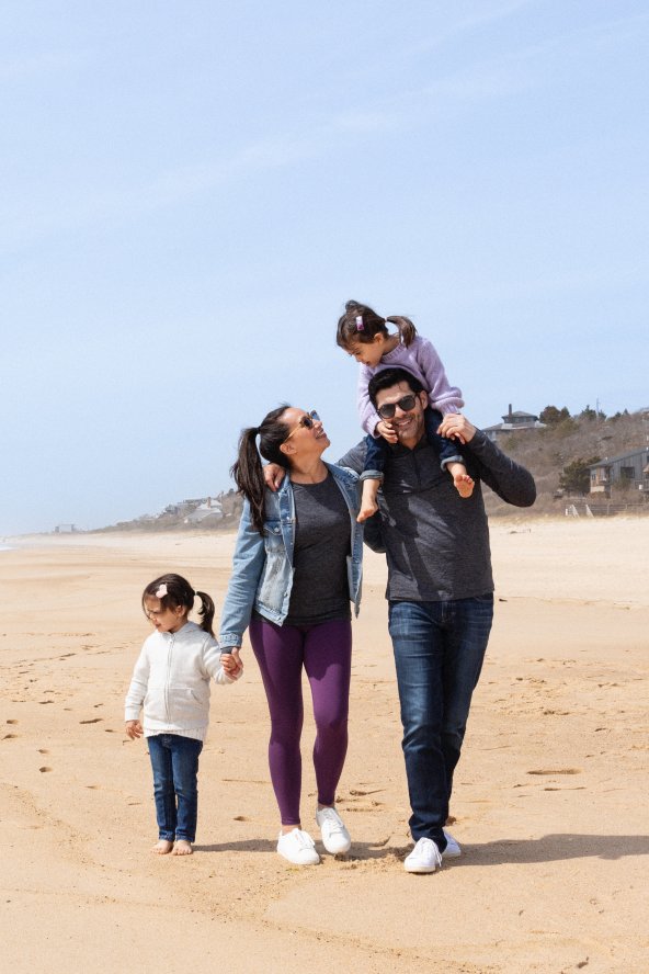 Family walking along a sandy beach, with the ocean on their left. The man carries a child on his shoulders, while another child walks beside the woman.