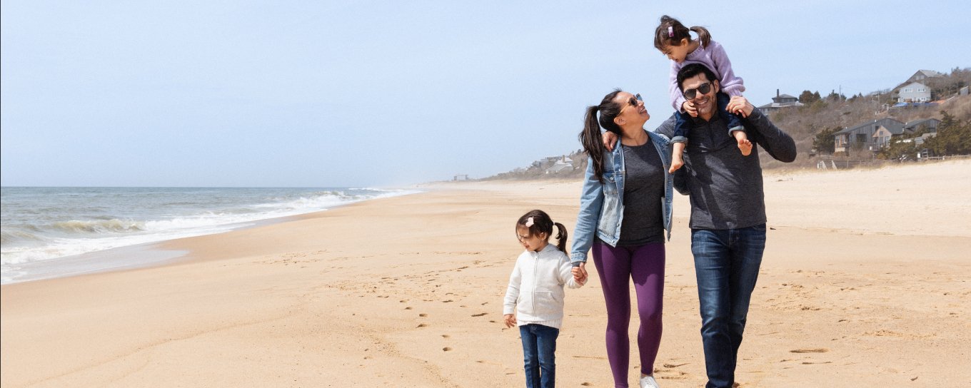 Family walking along a sandy beach, with the ocean on their left. The man carries a child on his shoulders, while another child walks beside the woman.