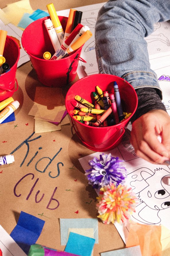 Children coloring at a table with red cups of markers and crayons, surrounded by paper and crafts. Brown paper reads "Kids Club."
