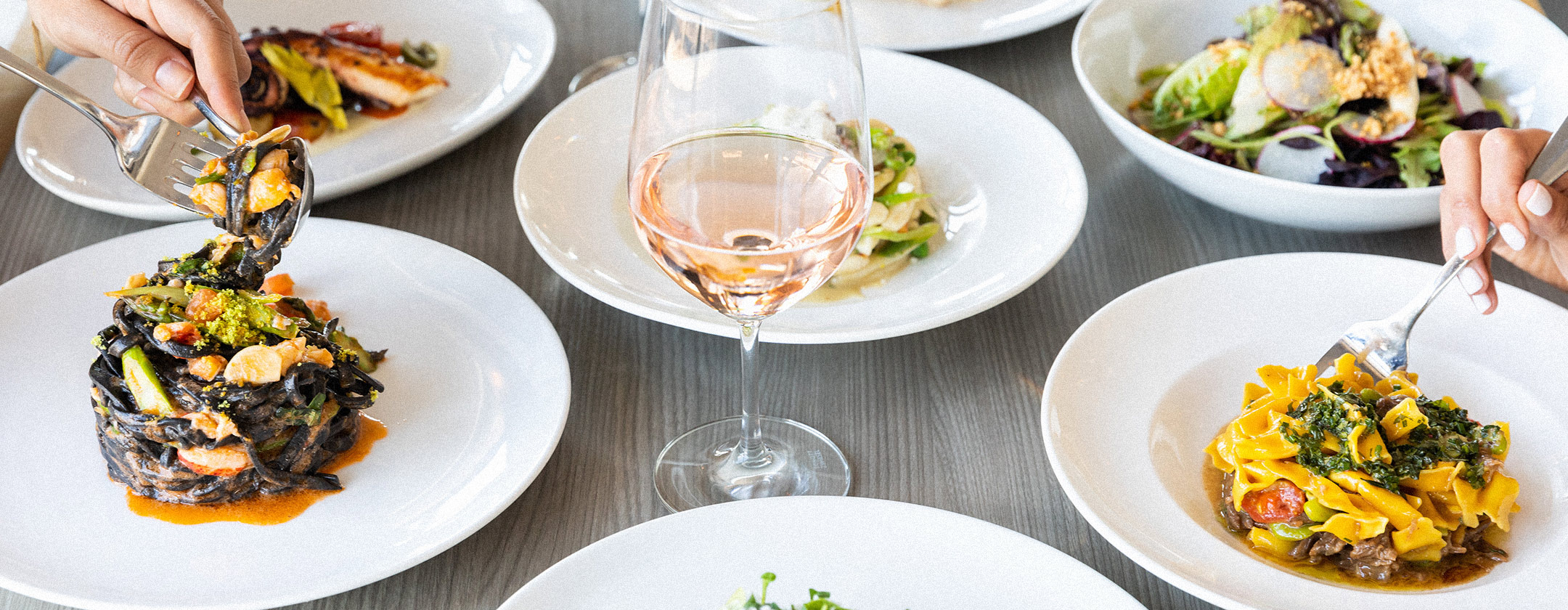 Hands serving pasta and salad dishes on white plates, with a glass of rosé wine in the center.