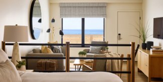 Cozy hotel room with ocean view, modern decor, a bed, sofa, TV, and large window. Neutral tones create a calm atmosphere.