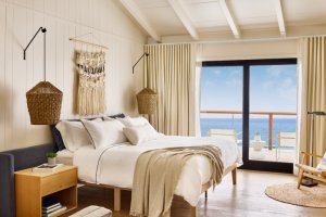 Modern bedroom with neutral tones, large bed, wicker accents, ocean view from sliding glass door, and natural light.