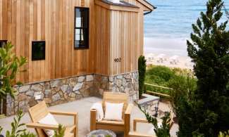 Wooden patio with cushioned chairs, lush green plants, and a sea view in the background.
