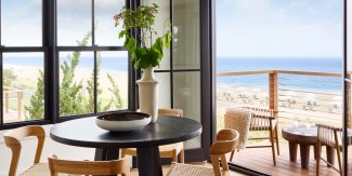 Cozy dining area overlooking a beach with blue ocean and clear sky, featuring a round table and wooden chairs by large glass windows and a balcony.
