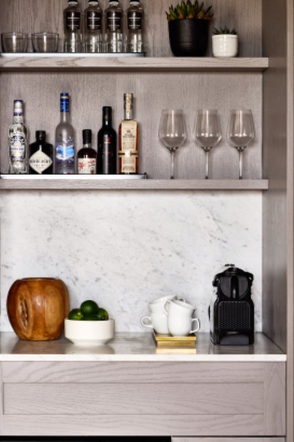 A mini bar with various liquor bottles, glasses, a bowl of limes, a wooden vessel, cups, a coffee machine, and a plant.