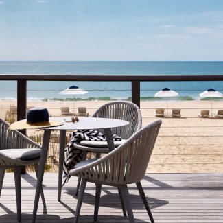 Two chairs and a table on a deck overlooking a sandy beach with white umbrellas and the ocean in the background.
