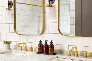 Elegant bathroom with two gold faucets, a white tiled wall, two mirrors, and three brown soap dispensers on a marble countertop.