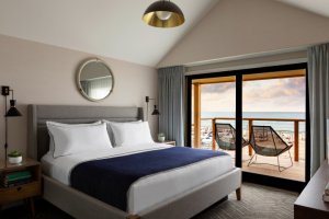 Cozy hotel room with a king bed, navy blanket, bedside tables, and a sliding door leading to a balcony with ocean view and two chairs.
