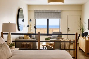 Cozy bedroom with a large bed, sofa, and decorative plants. A window reveals an ocean view, with a round mirror and modern lamps accenting the space.
