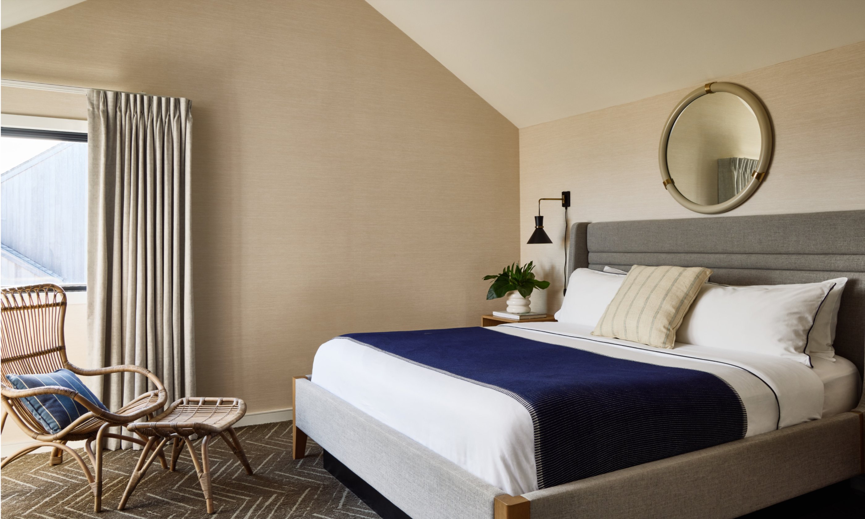 Contemporary bedroom with sloped ceiling, featuring a gray bed, navy blanket, round mirror, and rattan chair by a window with light curtains.