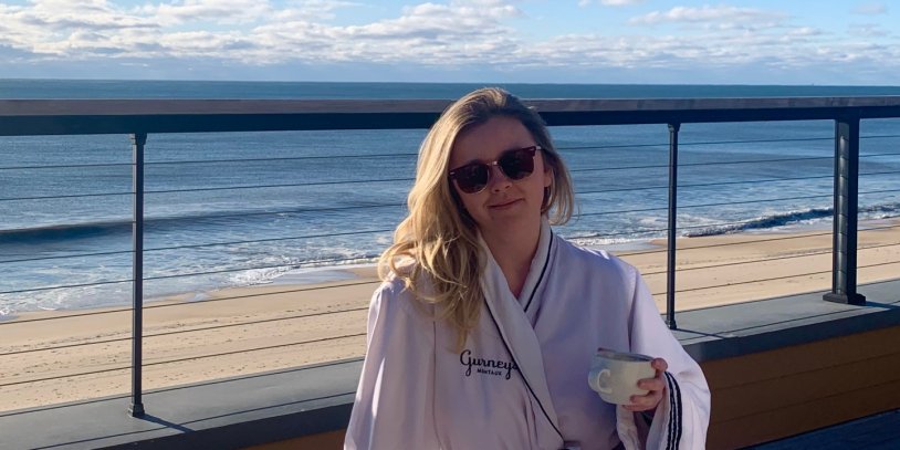 Woman in sunglasses and robe holding a cup, standing on a deck overlooking a calm ocean under a cloudy sky.