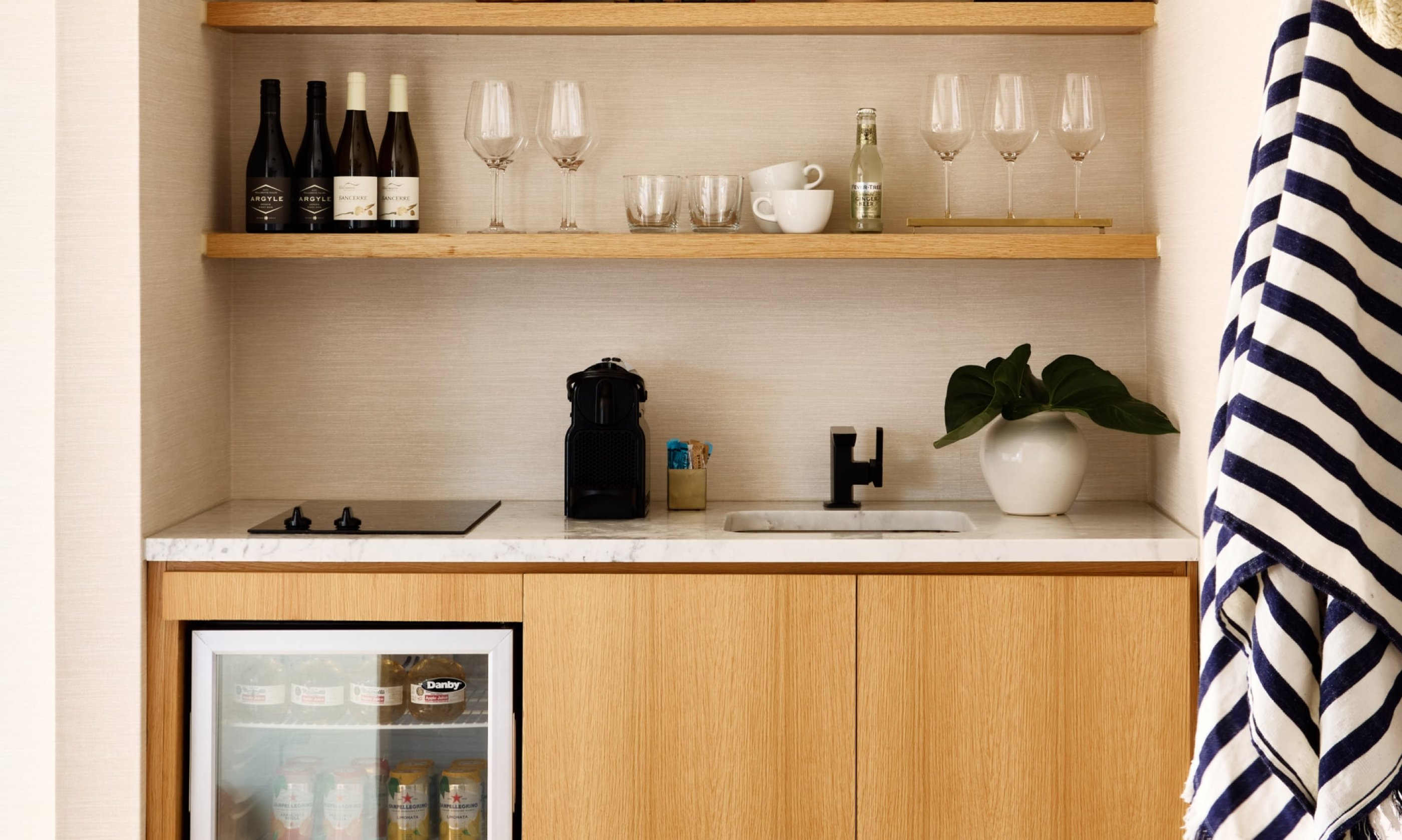 Kitchenette with wooden cabinets, a mini-fridge, wine bottles, glasses, a coffee maker, and a plant. Striped cloth hanging on the side.