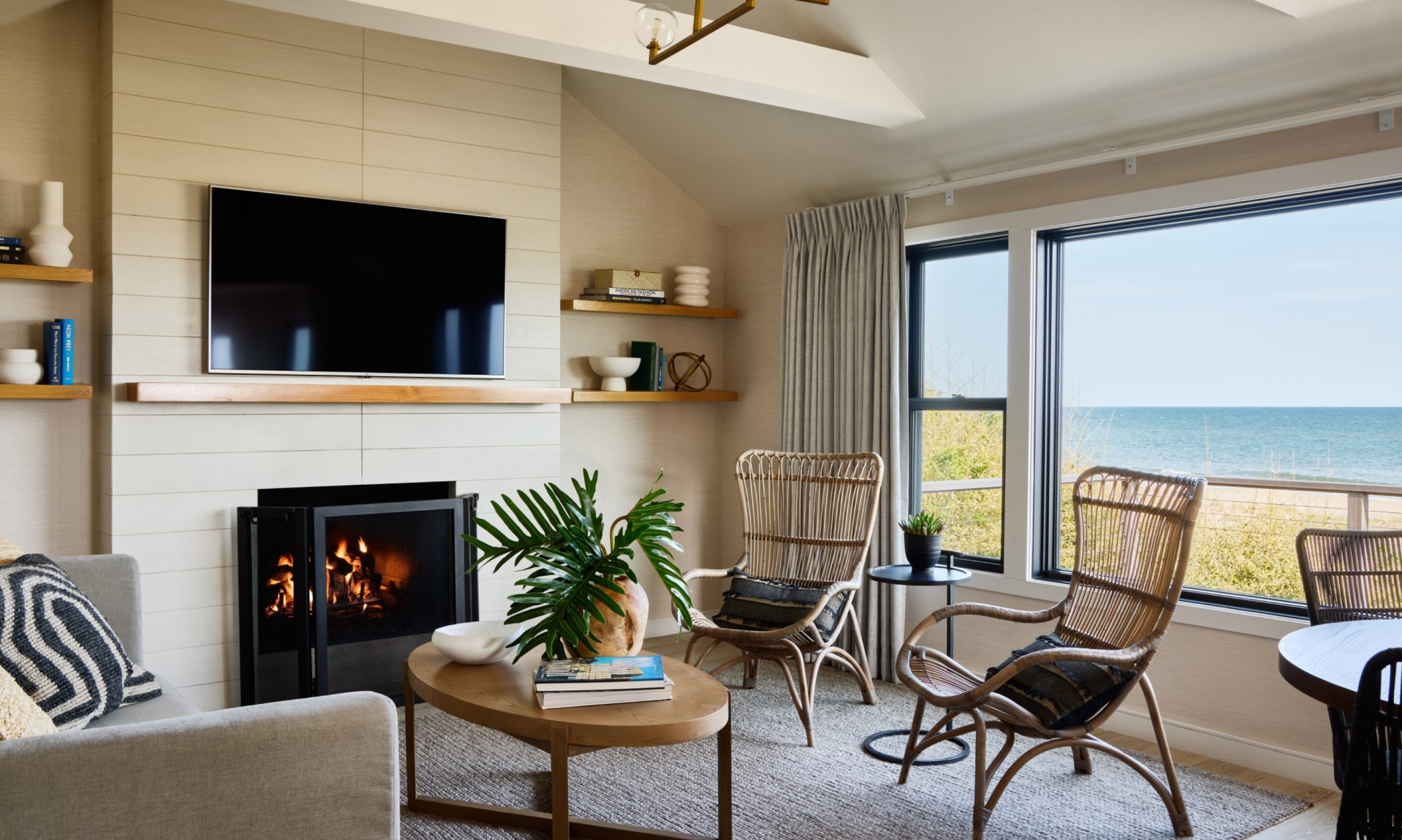 Cozy living room with a fireplace, large windows showing an ocean view, wicker chairs, and a neutral color palette.