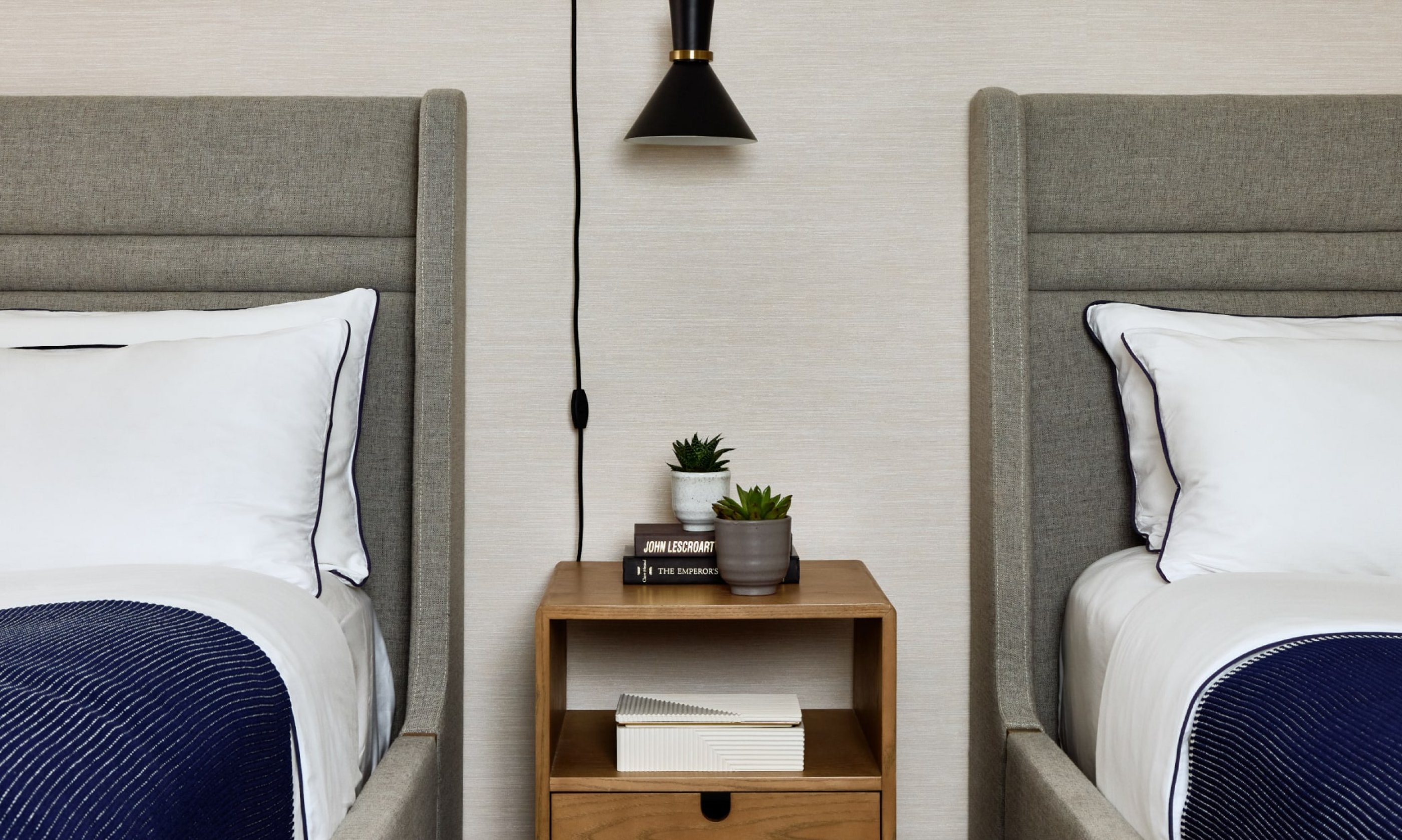 Two beds with grey headboards, a wooden nightstand between them with books and plants, and a black wall lamp above.
