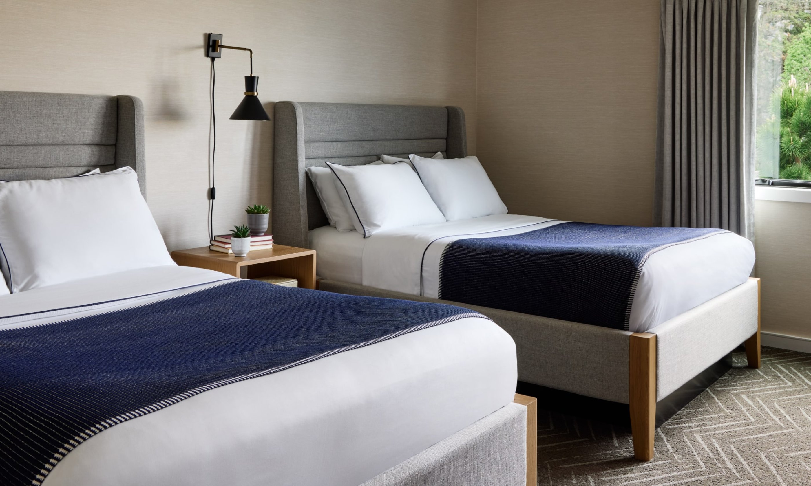 Two neatly made beds with gray headboards, white bedding, and navy throws in a hotel room with a window and soft lighting.
