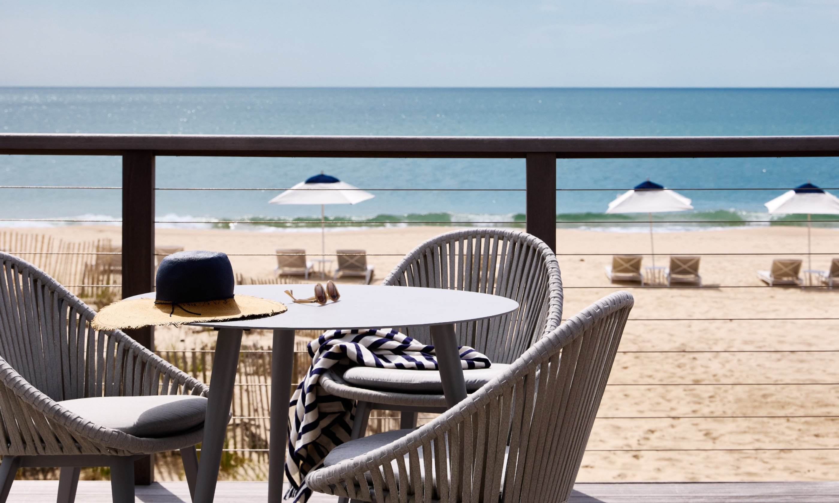Beachfront patio with two chairs, a table with a hat and scarf, overlooking sand, sea, and several beach umbrellas.