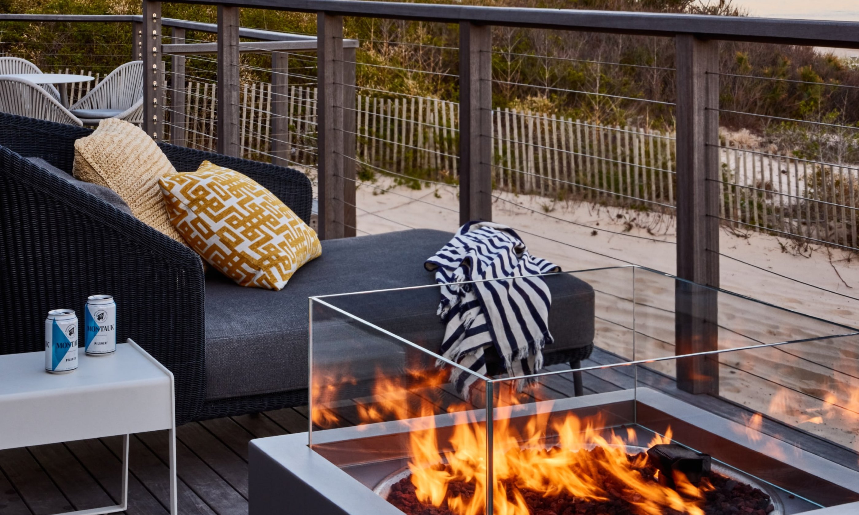 Modern patio with a gray sofa, yellow and white pillows, a glass fire pit, and a table with drinks, overlooking a sandy beach and wooden fence.