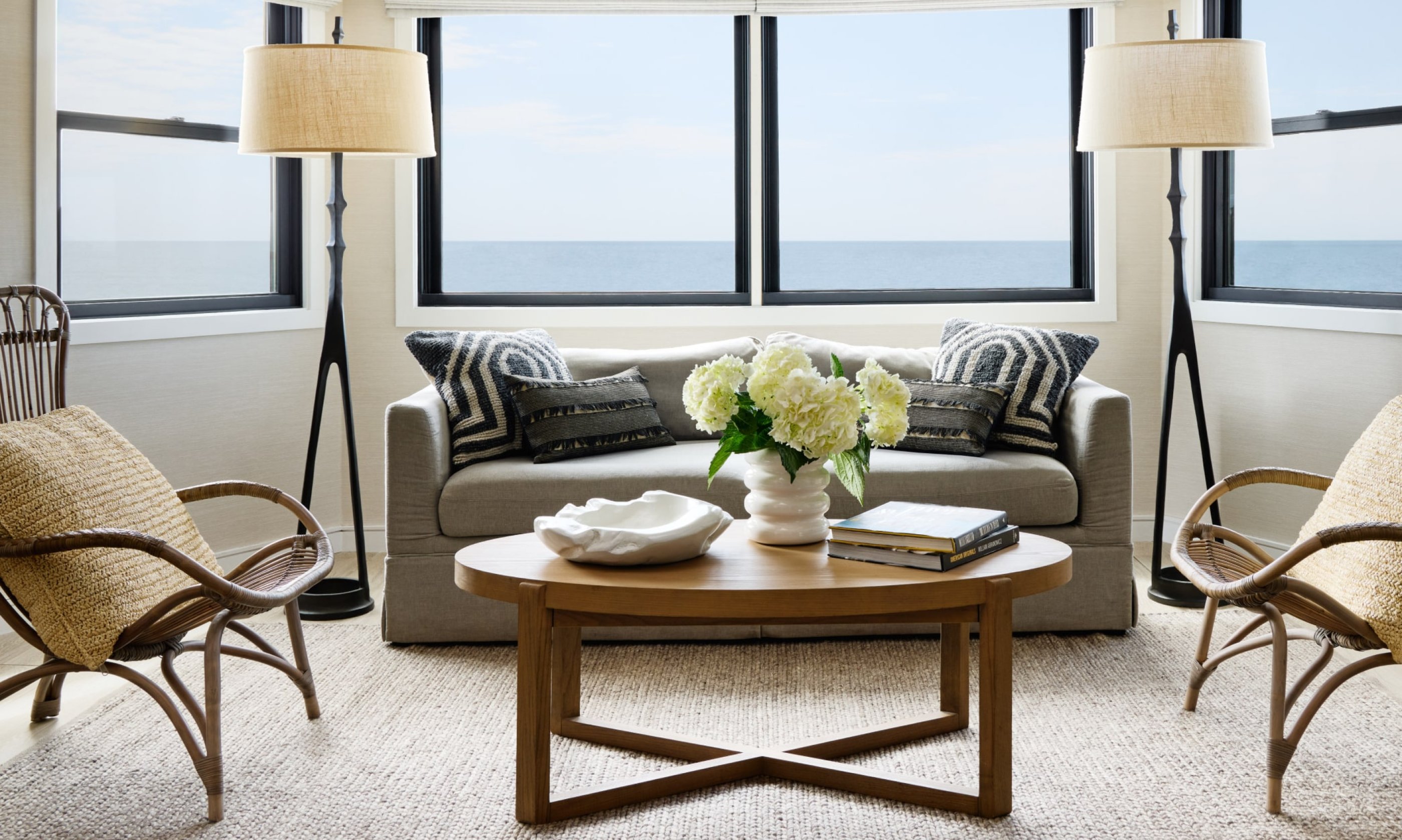 Cozy living room with a sea view, featuring a gray sofa, wooden table, and lamps. White flowers on the table, sand-colored chairs, and pillows.