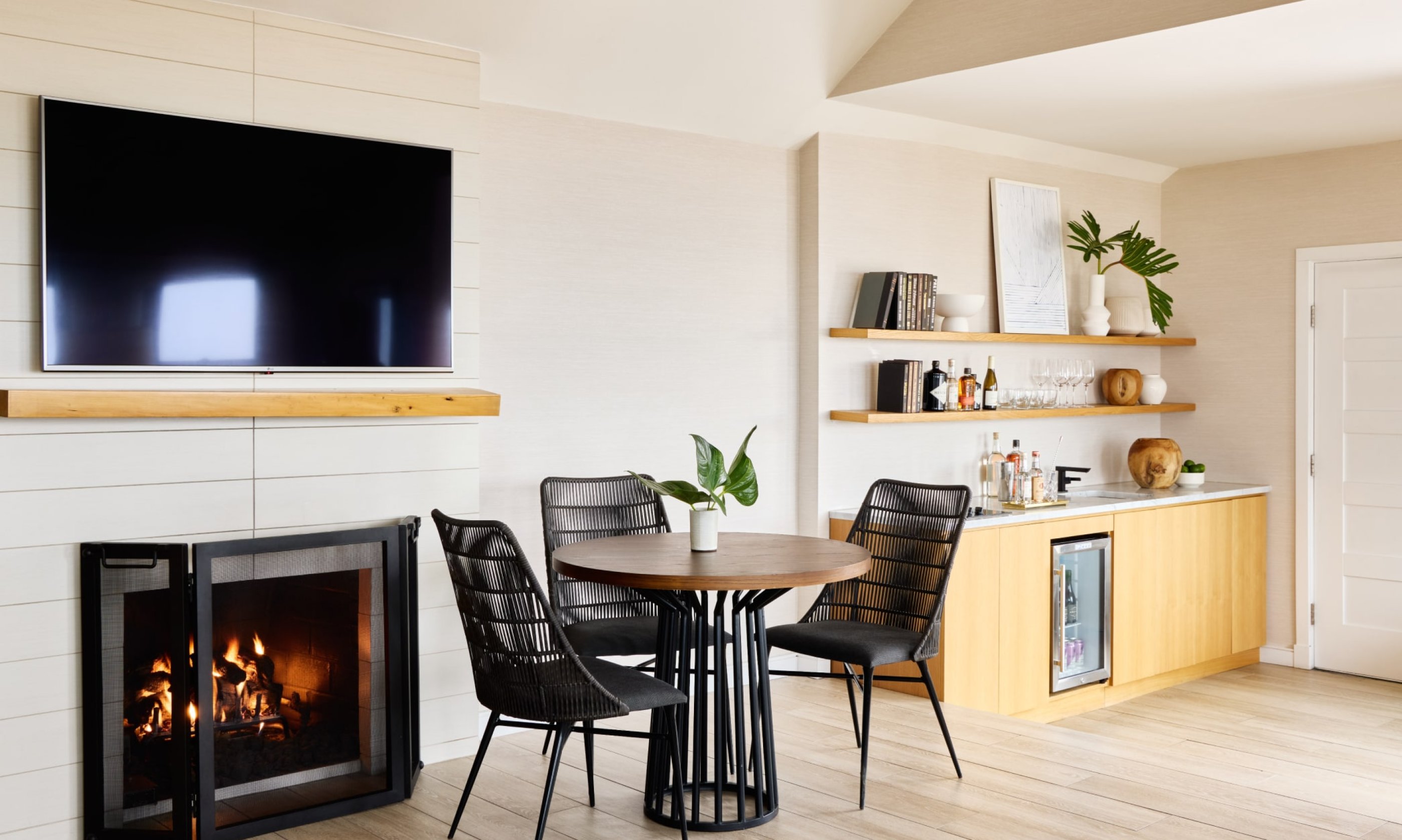 Cozy living area with a fireplace, TV, round table, and chairs. Light wood flooring and built-in shelves with decor on the right.