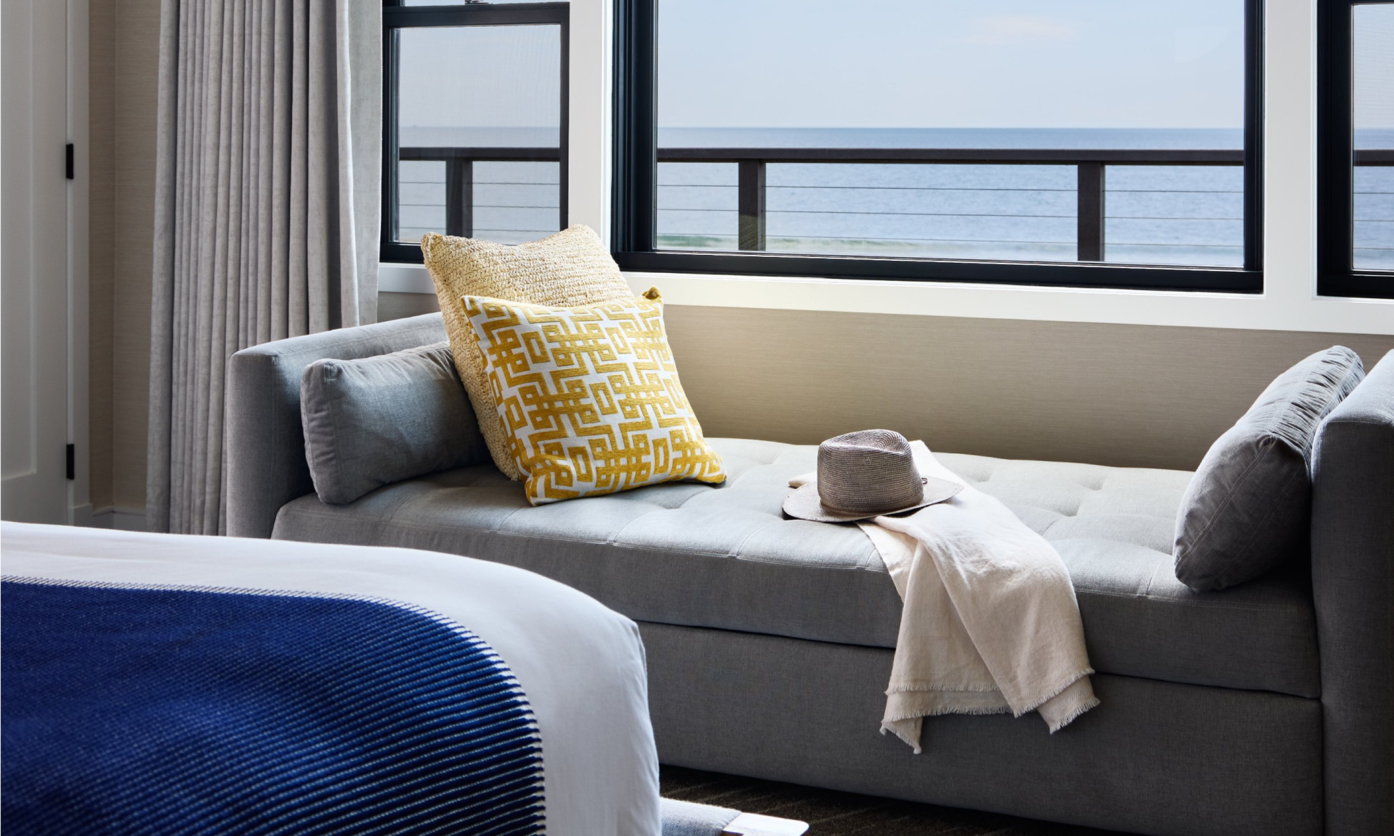 Cozy gray chaise lounge by window, ocean view. Yellow pillow, hat, and cream blanket on chaise. Blue blanket partially visible on bed.
