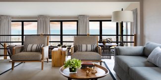 Cozy living room with a sea view, featuring two armchairs, a gray sofa, and tables with a plant and books. Large windows let in natural light.