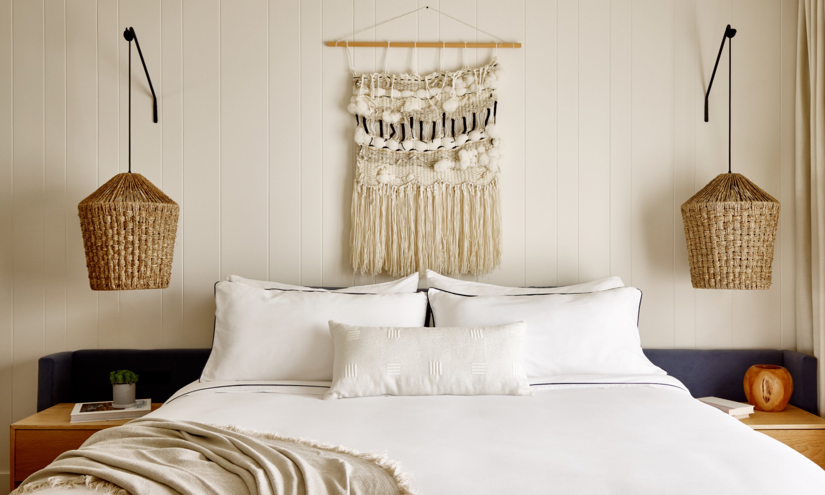 Cozy bedroom with a white bed, macramé wall hanging, and wicker hanging lamps. Neutral tones create a calm, minimalist look.