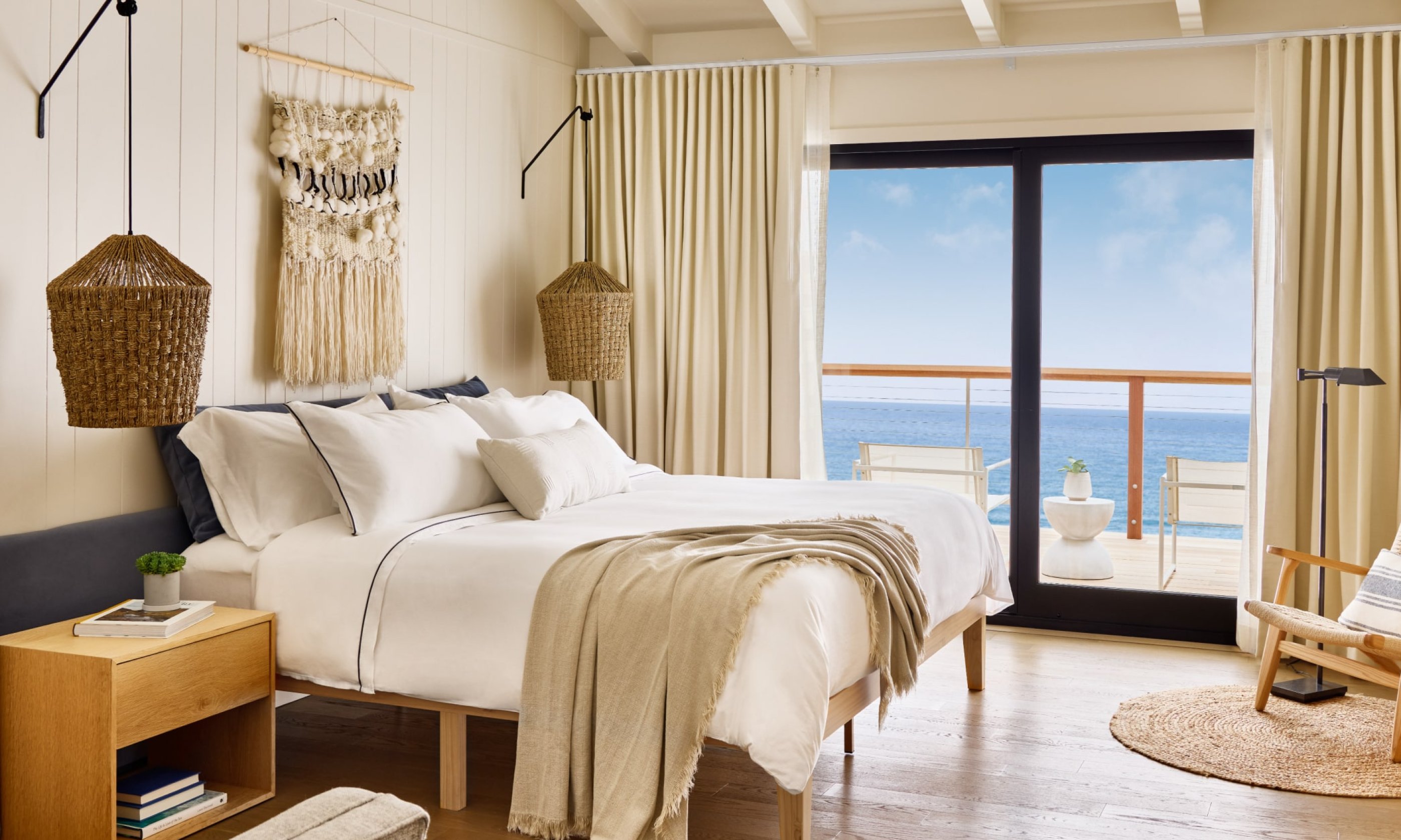 Sunny bedroom with ocean view, featuring a bed with white linens, a chair, and door leading to a balcony. Beige curtains and woven lamps add warmth.